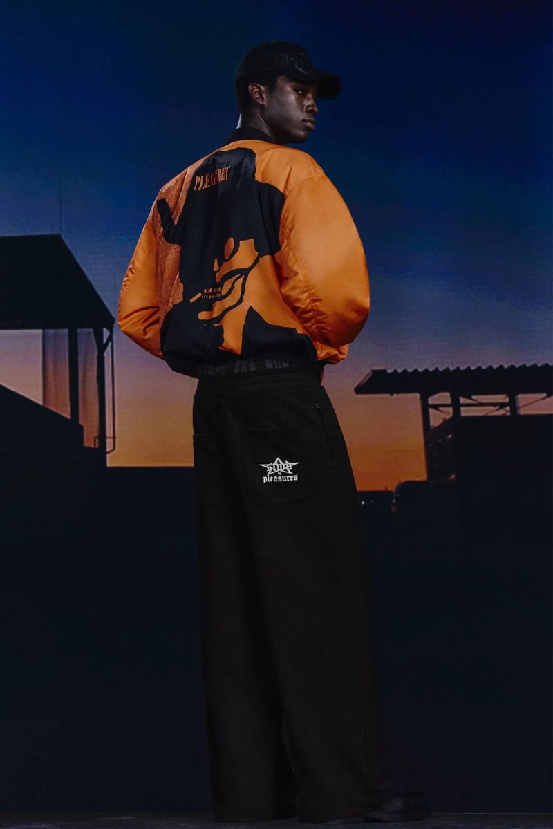 Man in orange bomber jacket and black pants stands outdoors at dusk, showcasing the garment's design and style.