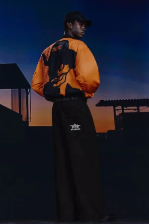 Man in orange bomber jacket and black pants stands outdoors at dusk, showcasing the garment's design and style.