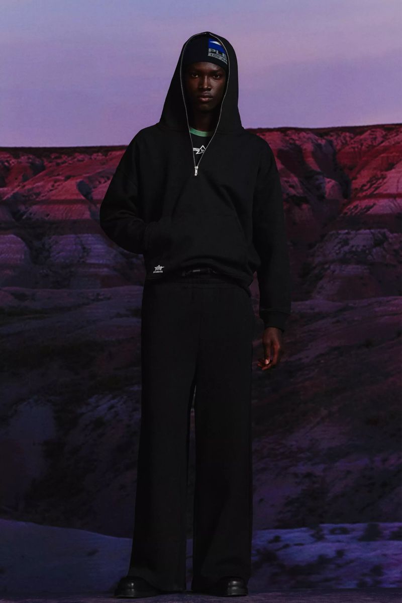 Full-length portrait of a Black male fashion model in a black hoodie and wide-leg pants, posed against a backdrop of a desert canyon.