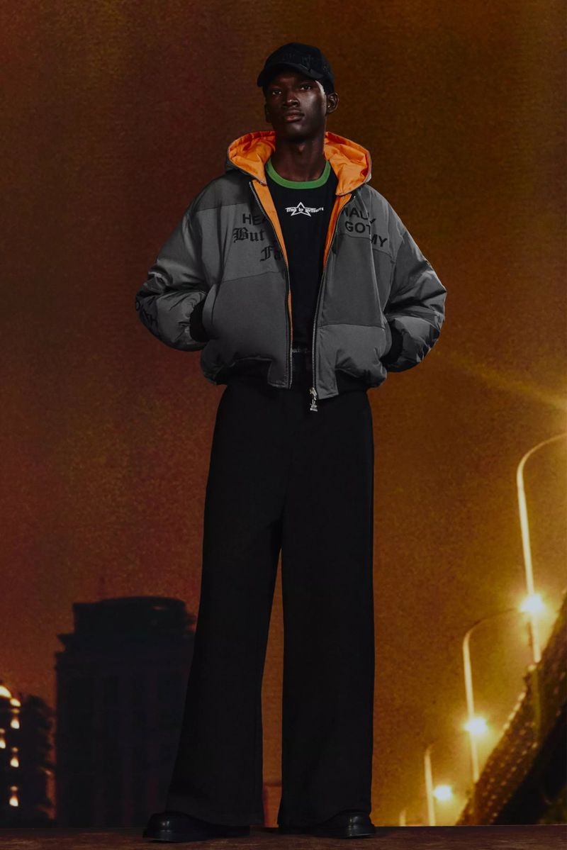 An African American male model stands in a gray hooded puffer jacket and black pants against a city backdrop, showcasing fashion apparel.