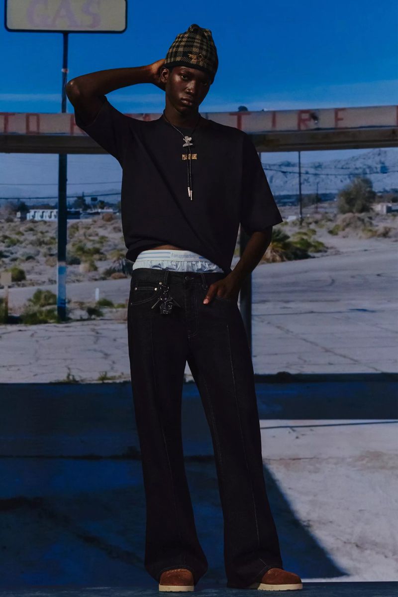 An adult Black male fashion model poses in a T-shirt, jeans, hat, and jewelry, standing in front of an abandoned structure in a desert setting.