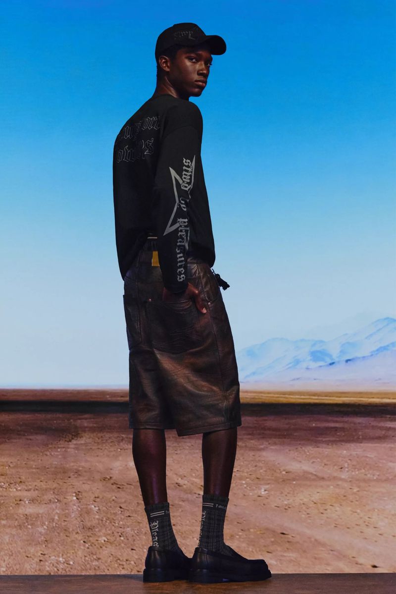A dark-skinned man in a black long-sleeved shirt, dark shorts, socks, and shoes poses against a desert backdrop with a blue sky gradient.