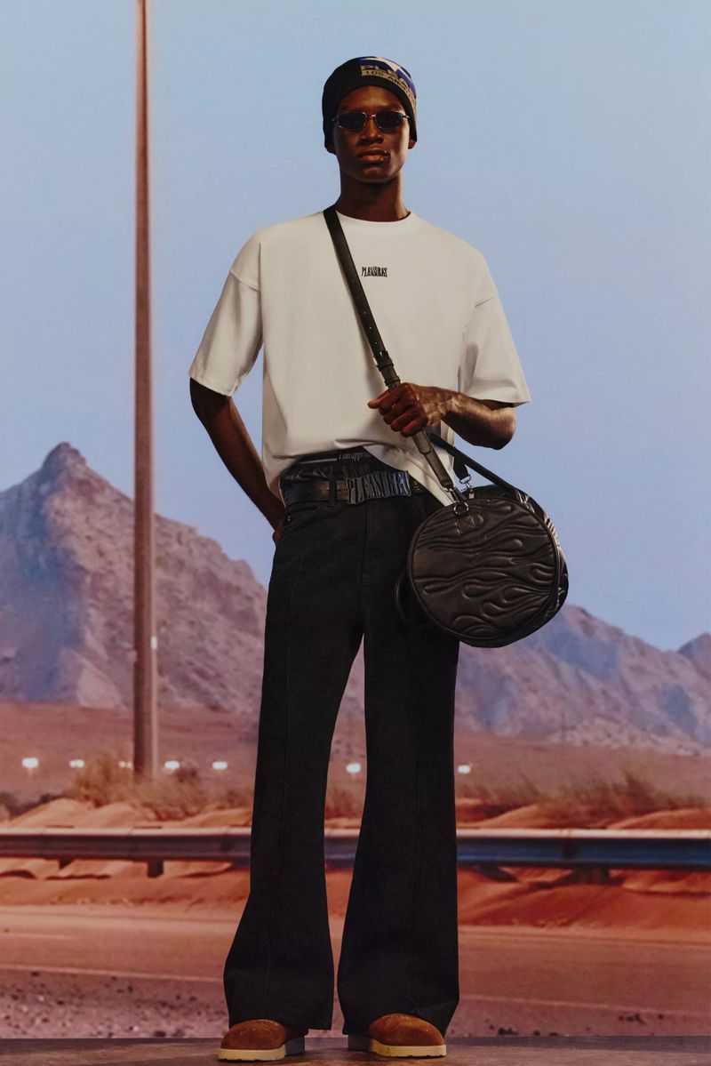 A Black male fashion model wears a white t-shirt, black pants, and a round handbag, standing in a desert-like setting with mountains in the background.