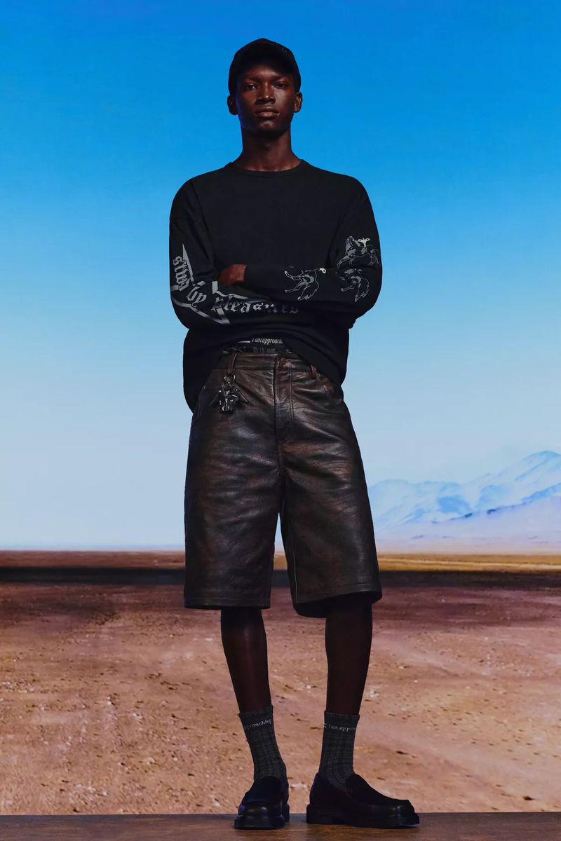 An adult male fashion model poses outdoors wearing a black long-sleeve shirt, brown leather shorts, socks, and shoes against a desert backdrop.