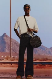 Model standing outdoors wearing a white t-shirt, dark pants, beanie, sunglasses, and carrying a round black crossbody bag.