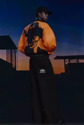 A Black man seen from behind wearing an oversized bright orange and black bomber jacket with wide-leg black pants at dusk.