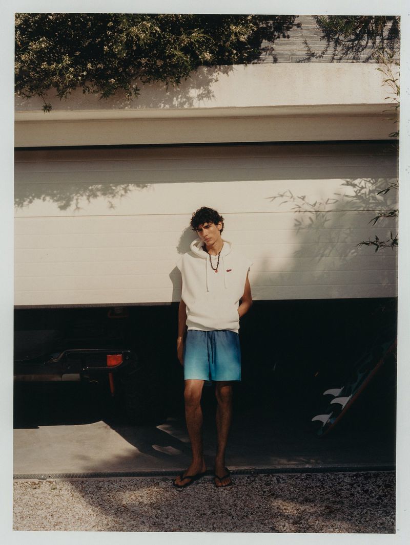 A young adult male wearing a sleeveless hoodie and blue shorts stands in front of a garage door with green foliage visible above.