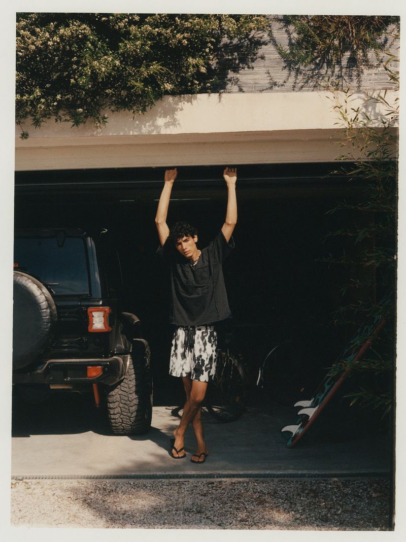 Young man in casual attire stands in a driveway in front of a Jeep and garage, with lush greenery above. Summer, outdoor setting.