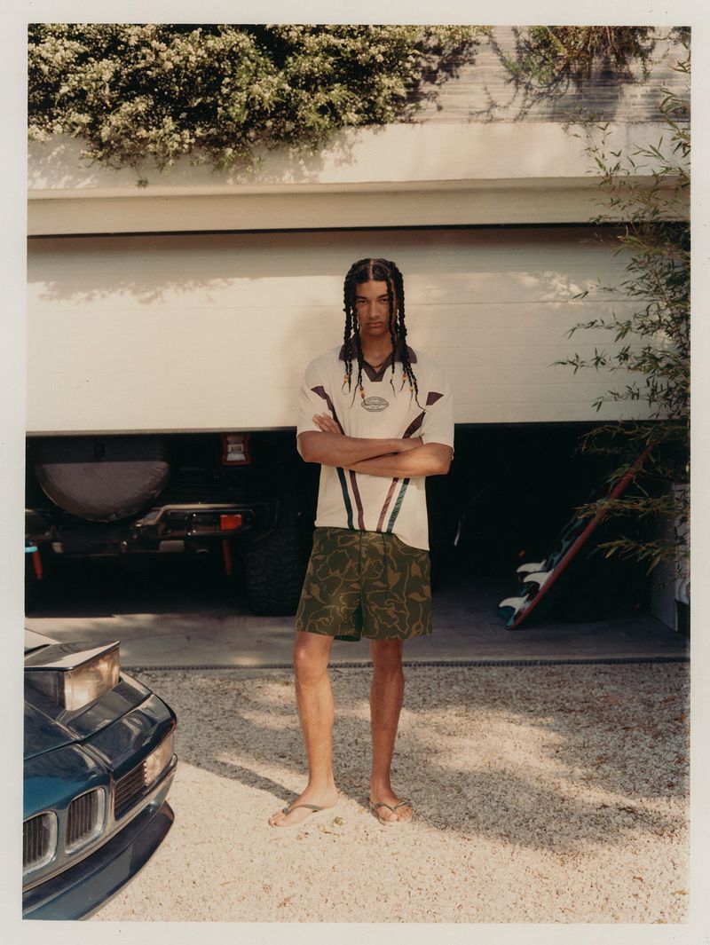 An adult male stands in front of a garage, wearing a cream and brown t-shirt with leaf-patterned shorts and sandals on a gravel driveway.