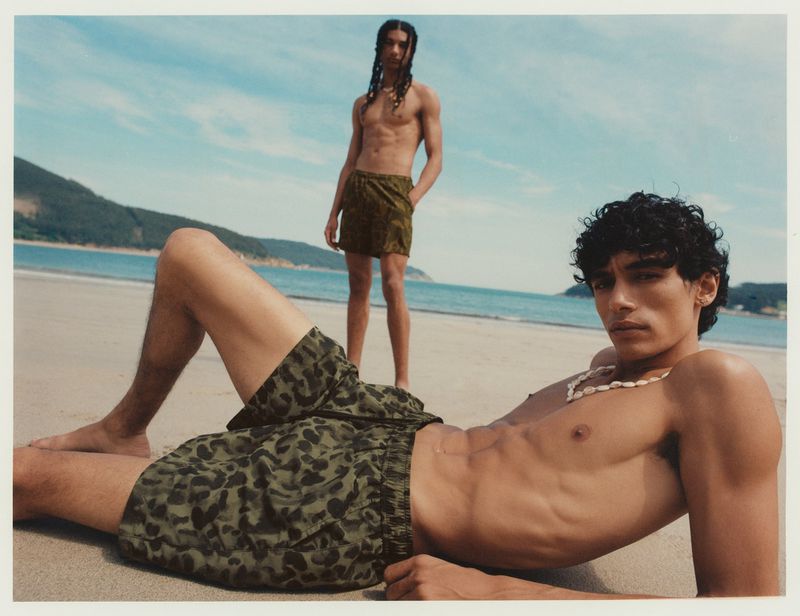 Two athletic adult males pose on a sandy beach with patterned swim trunks, necklaces, and a backdrop of ocean, sky, and distant hills.