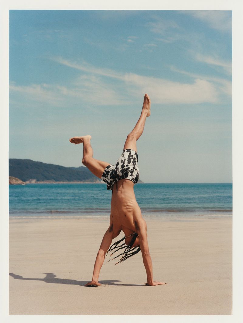 Shirtless man in patterned shorts doing a handstand on a sandy beach with a blue sea and sky in the background on a sunny day.