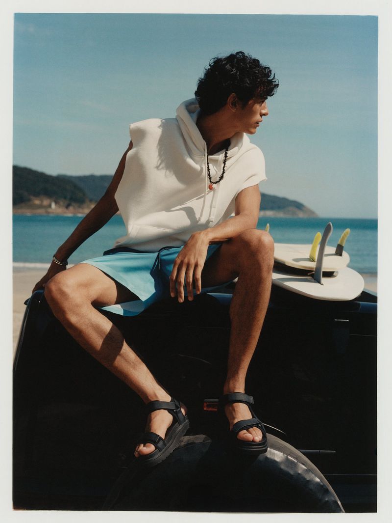 Man in hoodie and shorts sits on a car at the beach with a surfboard, set against the backdrop of the ocean and a sunny sky.