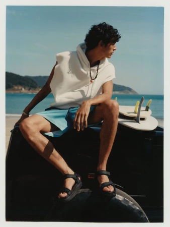 Man in hoodie and shorts sits on a car at the beach with a surfboard, set against the backdrop of the ocean and a sunny sky.