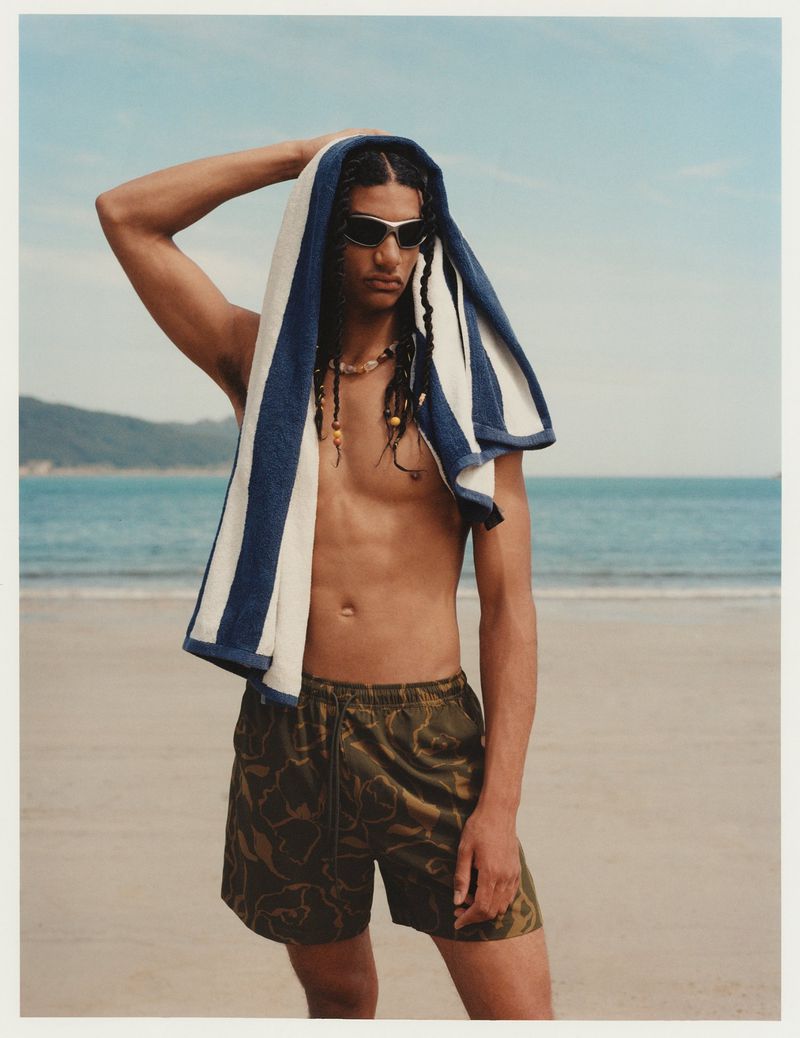 A tanned man with dreadlocks and sunglasses on a beach, draped with a striped towel, wears camouflage shorts against the coastal backdrop.