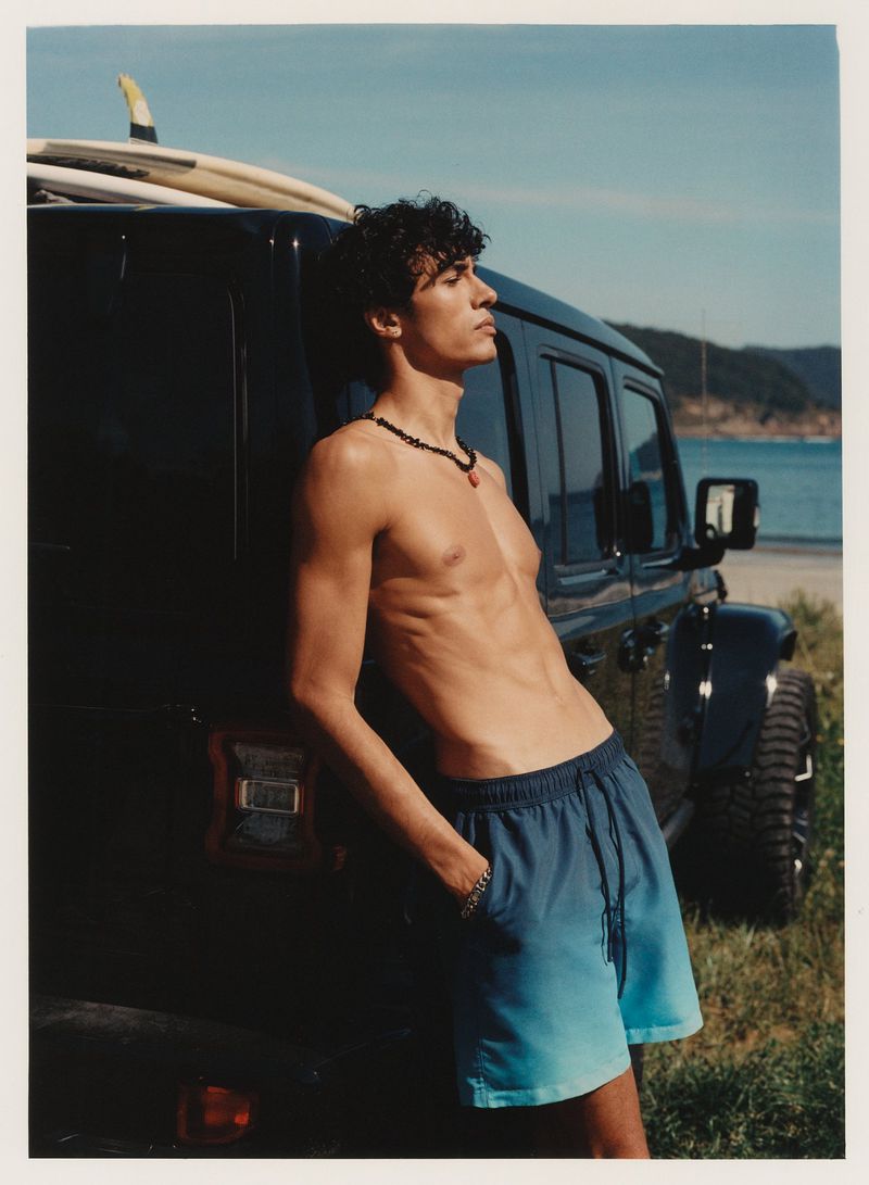Young man in swim trunks leans on a dark SUV with surfboards on top at a beach. A serene ocean and sky complete the scene.