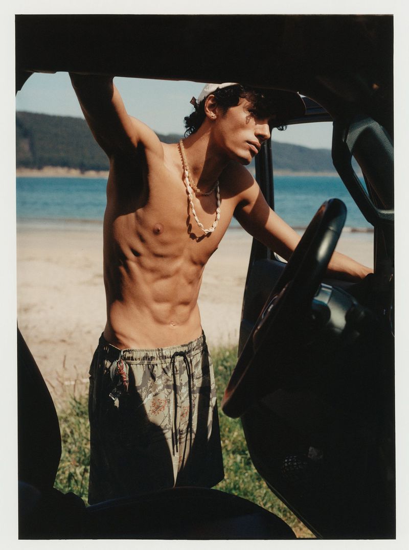 Shirtless man stands next to an SUV at the beach with mountains and water visible in the background on a sunny day.