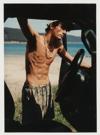 Shirtless man stands next to an SUV at the beach with mountains and water visible in the background on a sunny day.