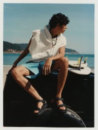 Man in a white sleeveless hoodie and blue shorts sitting on a truck bed with surfboards overlooking the ocean.