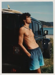 Shirtless young man leaning against a dark car by the coast wearing gradient blue swim trunks and a beaded necklace