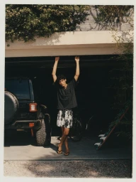 Model Zion standing under an open garage frame wearing an oversized black t-shirt and printed shorts next to a black vehicle.