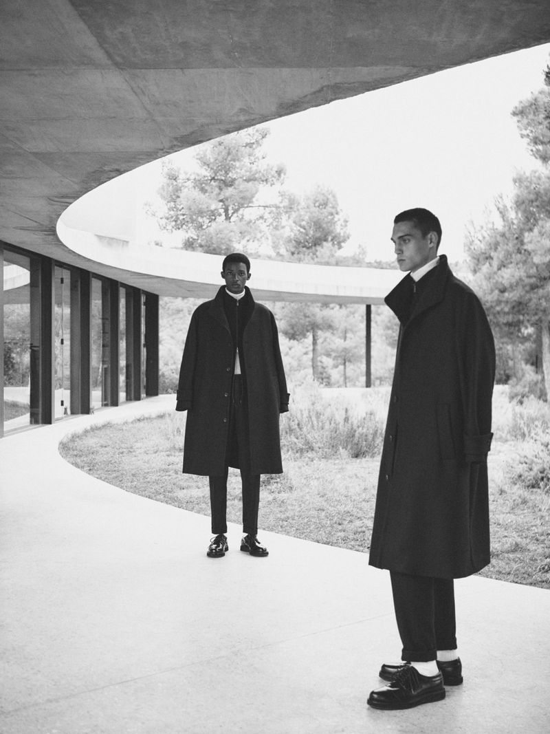 Two men in dark overcoats stand in a modern architectural setting with trees and light stone. Editorial fashion shot in black and white.