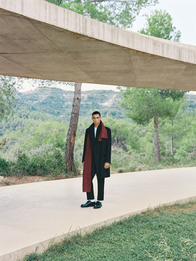 An adult male standing outdoors wearing a dark coat and scarf, posing underneath a concrete overhang with a forest in the background.