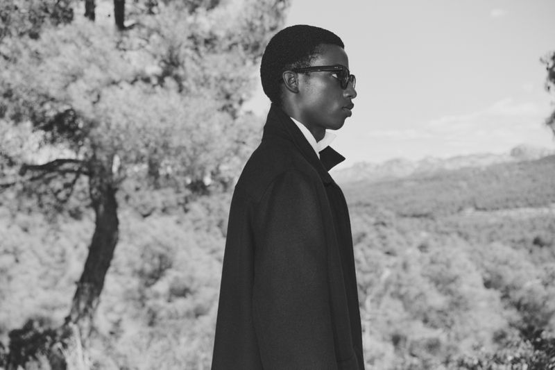 Monochrome portrait of a young African-American man in sunglasses and a coat, set against a blurred nature landscape backdrop.