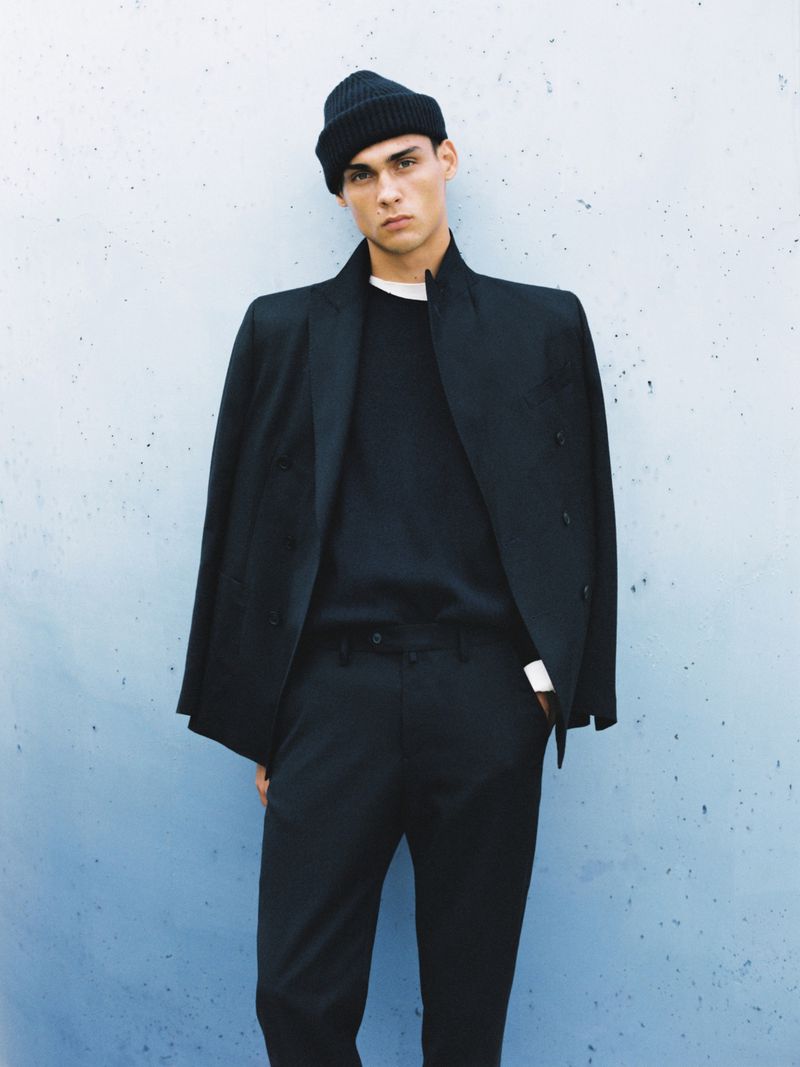 A young adult male in a black wool beanie, coat, pants, and sweater poses indoors against a textured grey-blue wall. Fashion shot, portrait.