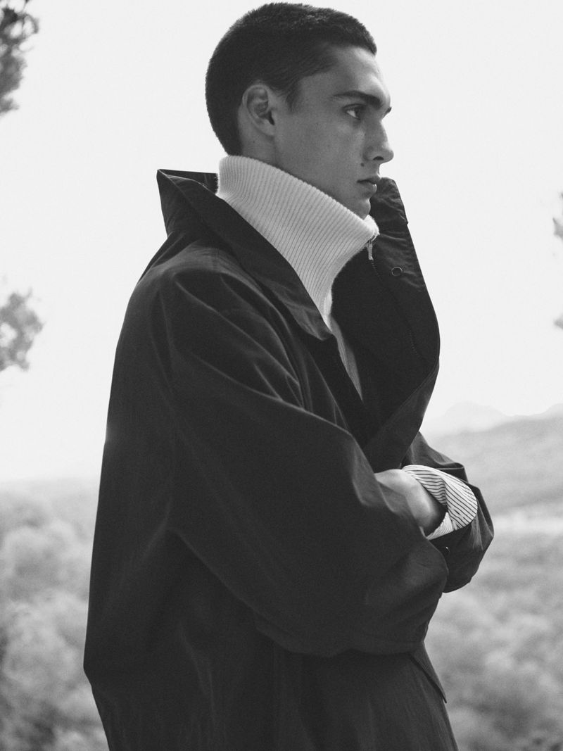 Monochrome portrait of a young adult male in a black jacket and white turtleneck, gazing to the side against a natural outdoor background.