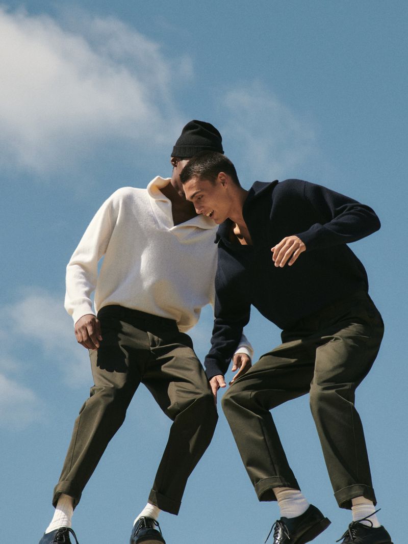Two young men in a fashionable clothing shot at a low angle, set against a blue sky with soft clouds, one in a black beanie.