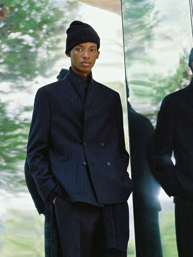 Studio shot of a Black male model in a pinstripe suit and beanie, reflected in a mirror, with a neutral expression and soft lighting.