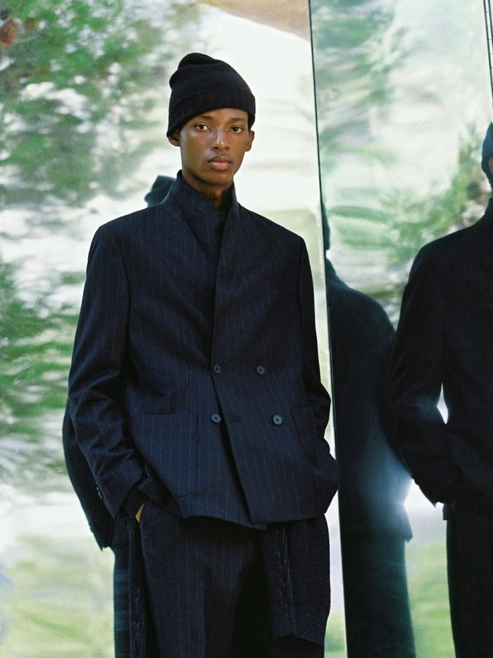Model wearing a dark pinstripe blazer and black beanie standing against a reflective glass surface