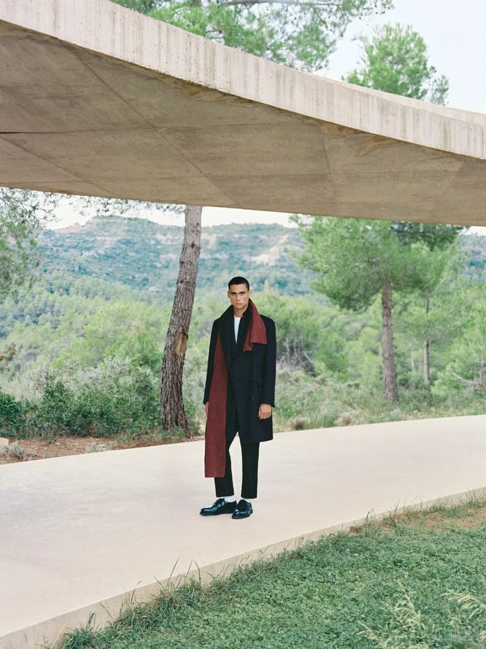 Male model wearing a dark coat and red scarf standing on a pathway under a concrete structure outdoors