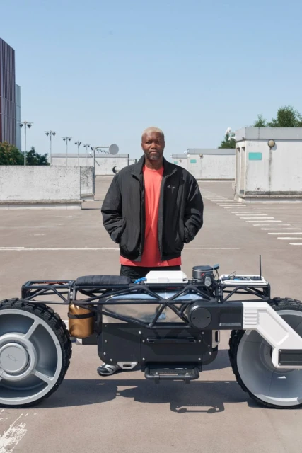 Man poses with robotic vehicle outdoors on a concrete rooftop with buildings and a clear blue sky in the background during the day.