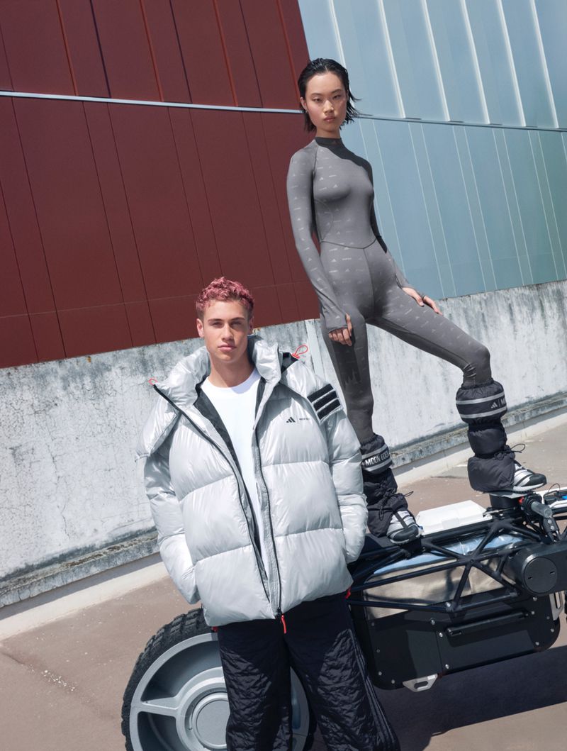 Two models pose outdoors in athletic wear by a futuristic vehicle. One wears a puffer jacket and the other is in a grey catsuit.