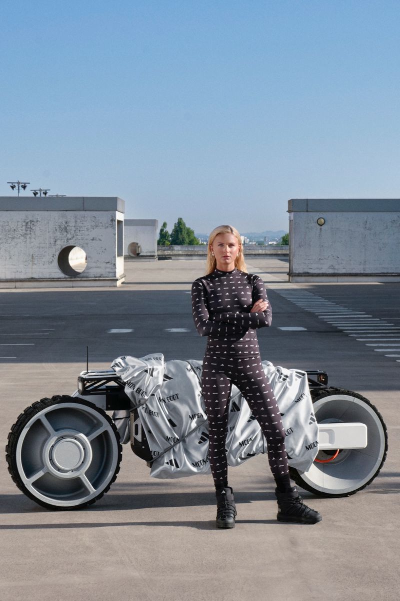 A woman in a patterned body suit stands in front of a covered motorbike on a rooftop with a minimalist architectural background on a sunny day.