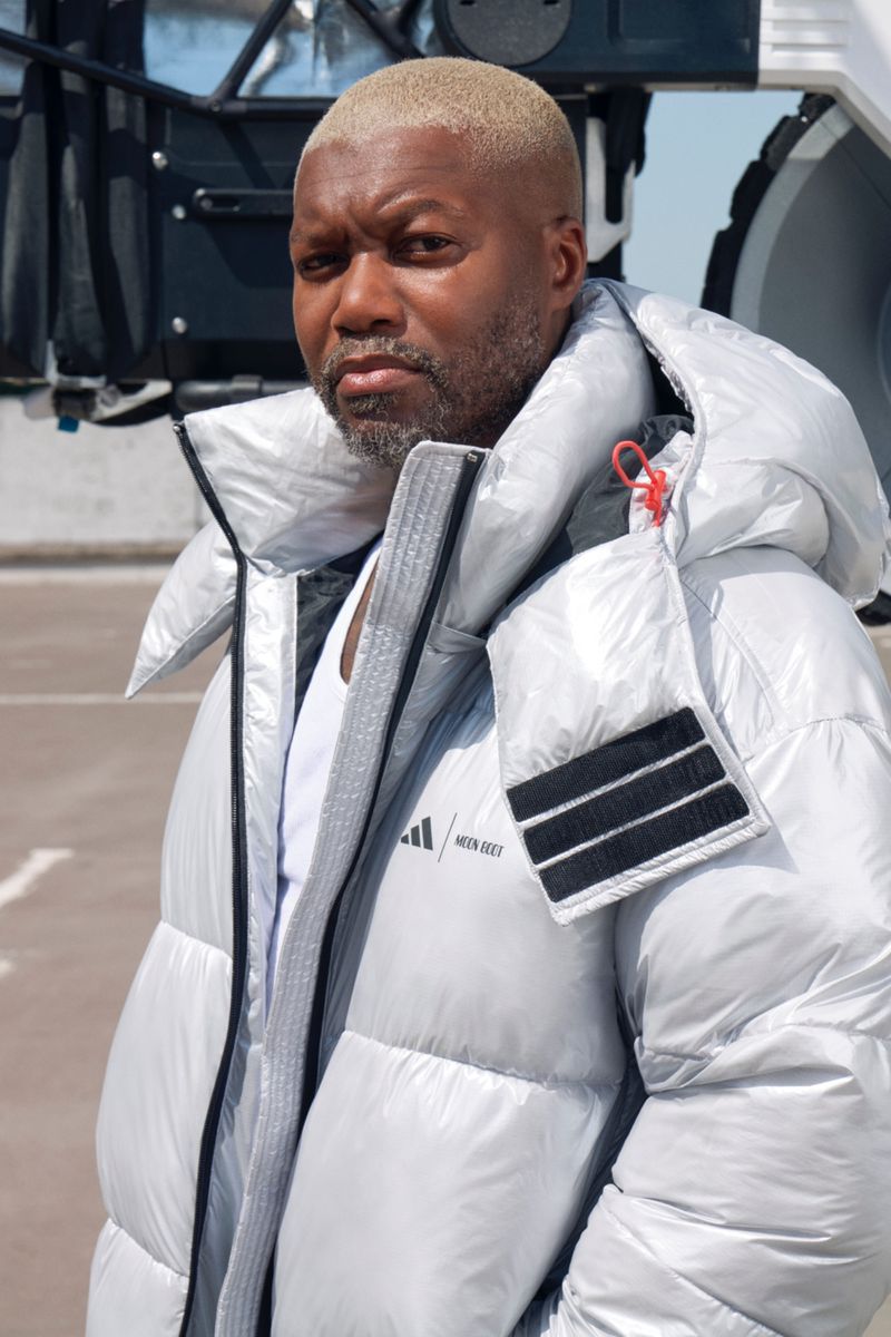 Close-up of an adult Black male wearing a silver puffer jacket and a white t-shirt outdoors during daylight, with a neutral expression.