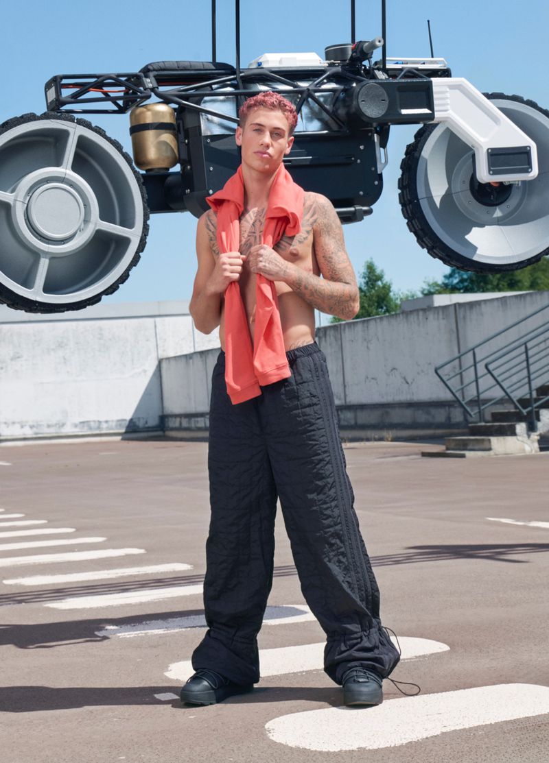 Adult male with pink hair poses with futuristic vehicle on concrete rooftop in black pants and coral towel against a blue sky.