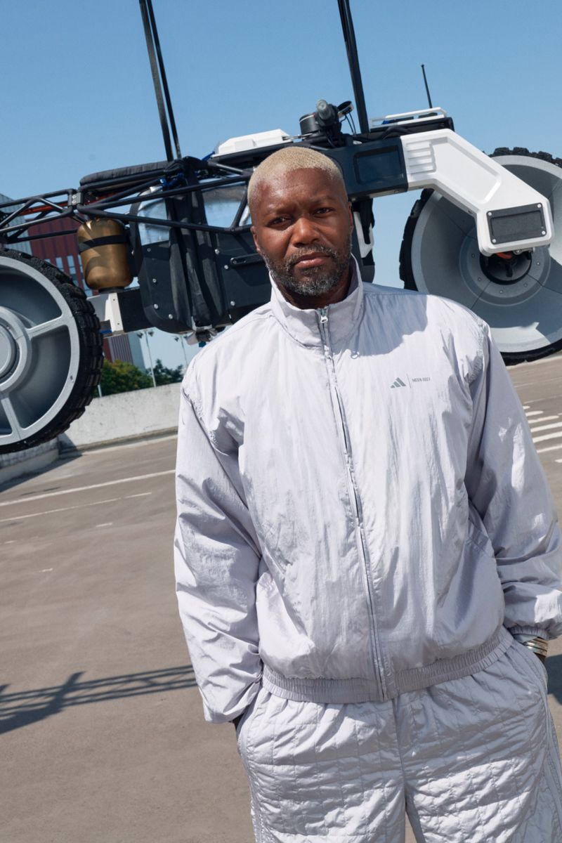 An adult male stands in front of a large, wheeled, mechanical structure wearing a matching gray tracksuit outdoors under a blue sky.