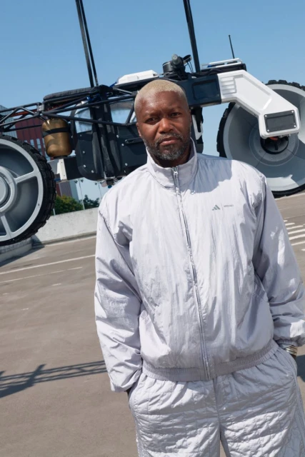 An adult male stands in front of a large, wheeled, mechanical structure wearing a matching gray tracksuit outdoors under a blue sky.