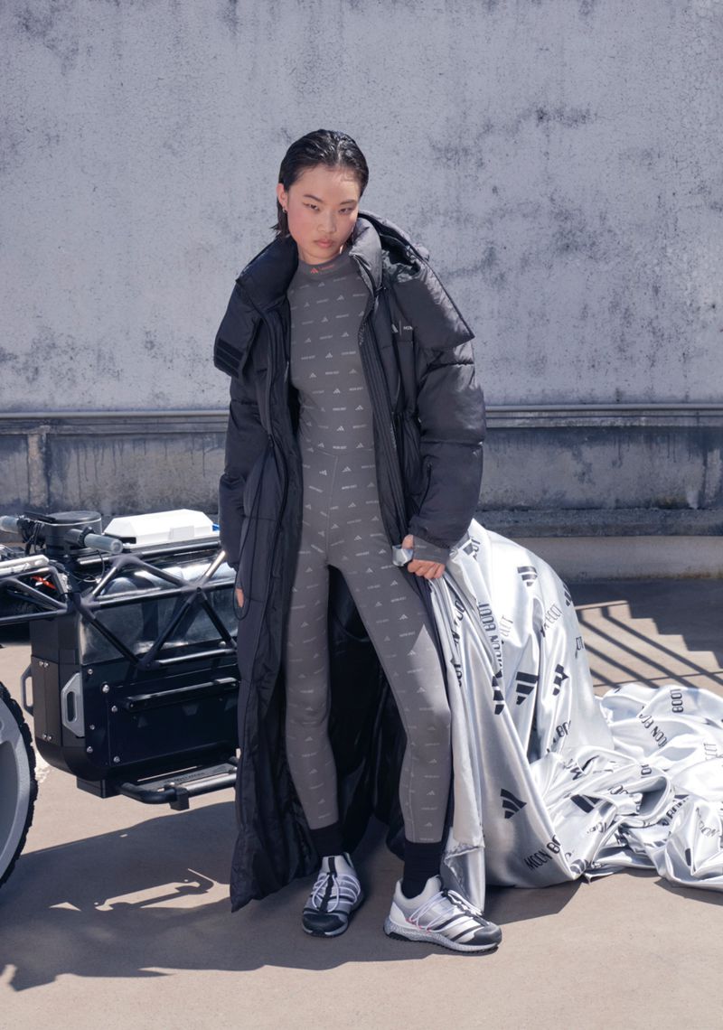 A woman models a grey patterned bodysuit and black puffer coat on a rooftop, showcasing fashion against a backdrop of silver bags and luggage.