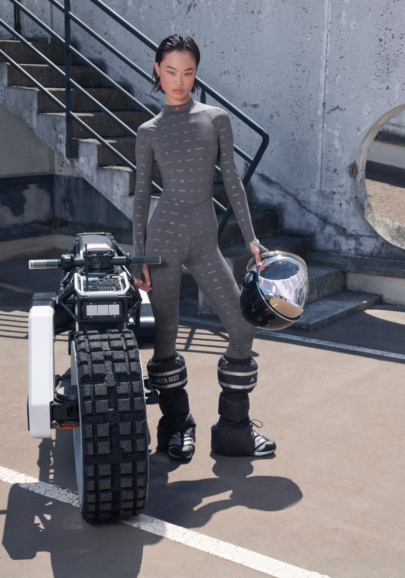 Stylish Asian model in grey catsuit poses with an electric motorbike on an urban rooftop, holding a black motorcycle helmet.
