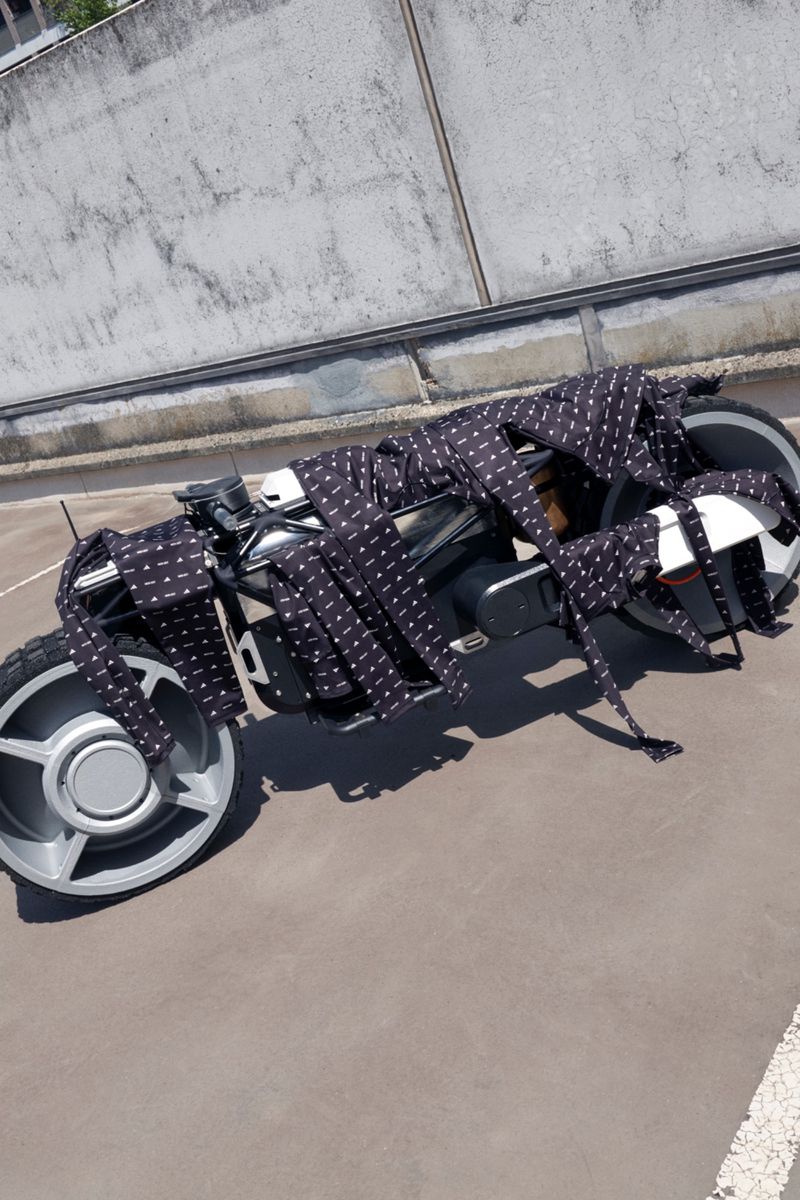 Futuristic motorcycle is decoratively draped with multiple neckties, displayed outdoors against a concrete background on a sunny day.