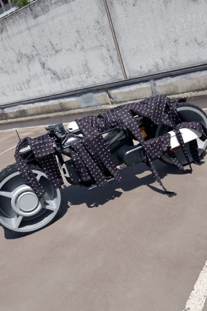 Futuristic motorcycle is decoratively draped with multiple neckties, displayed outdoors against a concrete background on a sunny day.