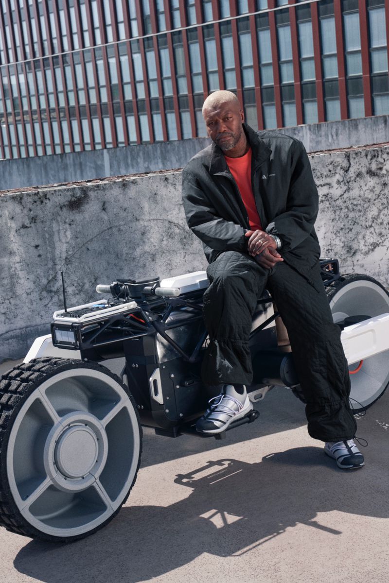 Bald African American man sits on an all-terrain vehicle against a building backdrop, wearing sportswear in an outdoor setting.