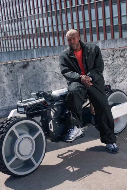Bald African American man sits on an all-terrain vehicle against a building backdrop, wearing sportswear in an outdoor setting.
