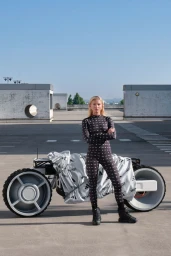 Model in a black printed catsuit standing in front of a futuristic concept motorcycle on an open concrete rooftop.