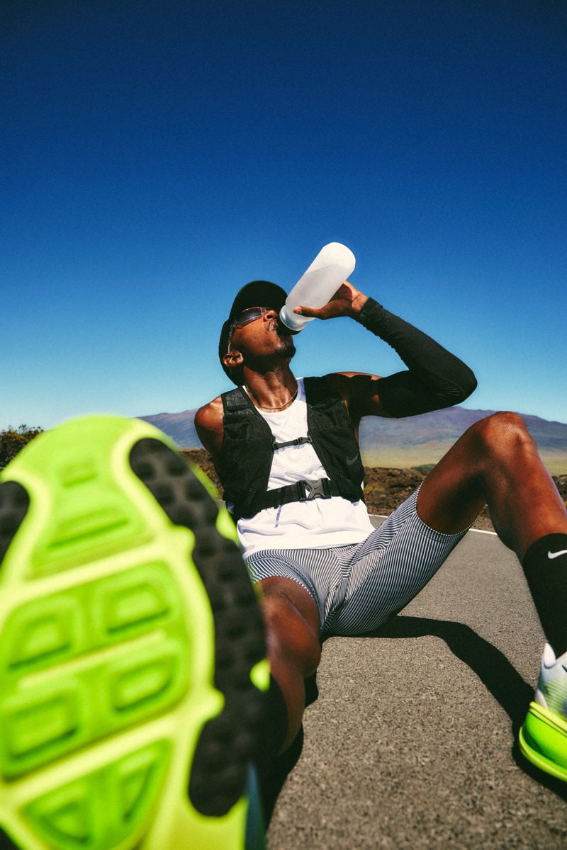 An African American man in sportswear drinks from a water bottle on a road, wearing a vest, sunglasses, and a hat, with a bright blue sky above.
