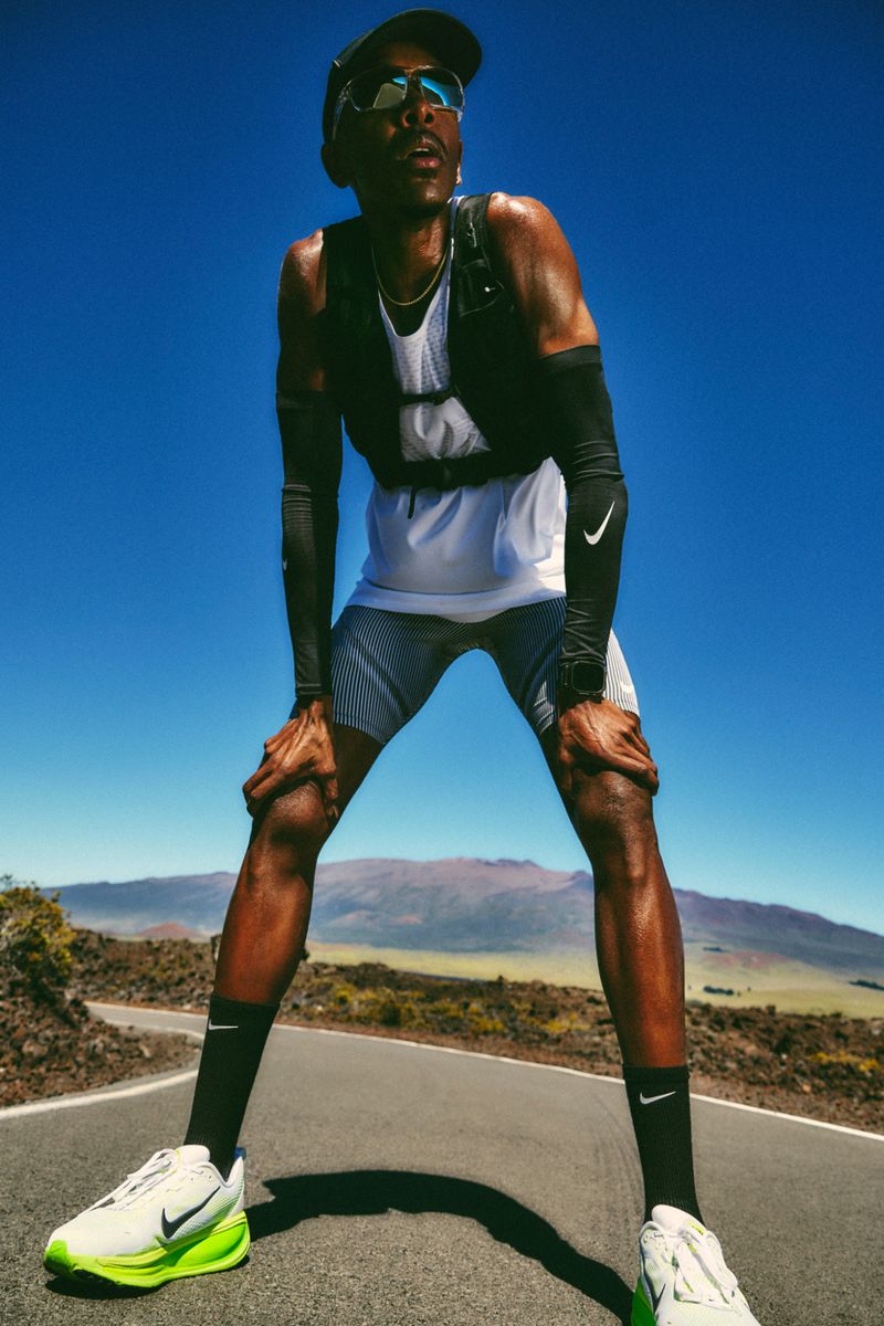 A male runner pauses on a road, hands on knees, wearing sportswear and sunglasses under a bright blue sky with a blurred landscape.