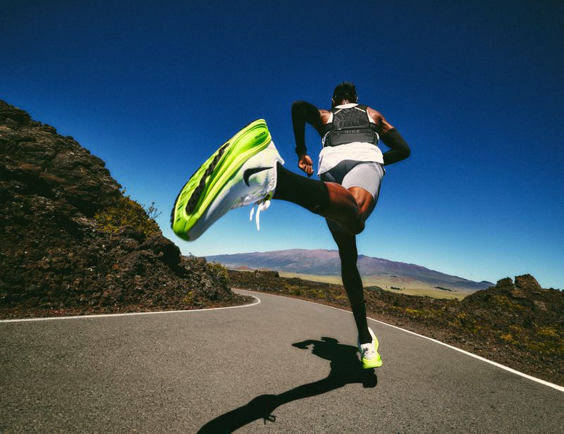 Athlete in motion on a road, neon running shoes in the air, set against a backdrop of a clear sky and distant mountain range.